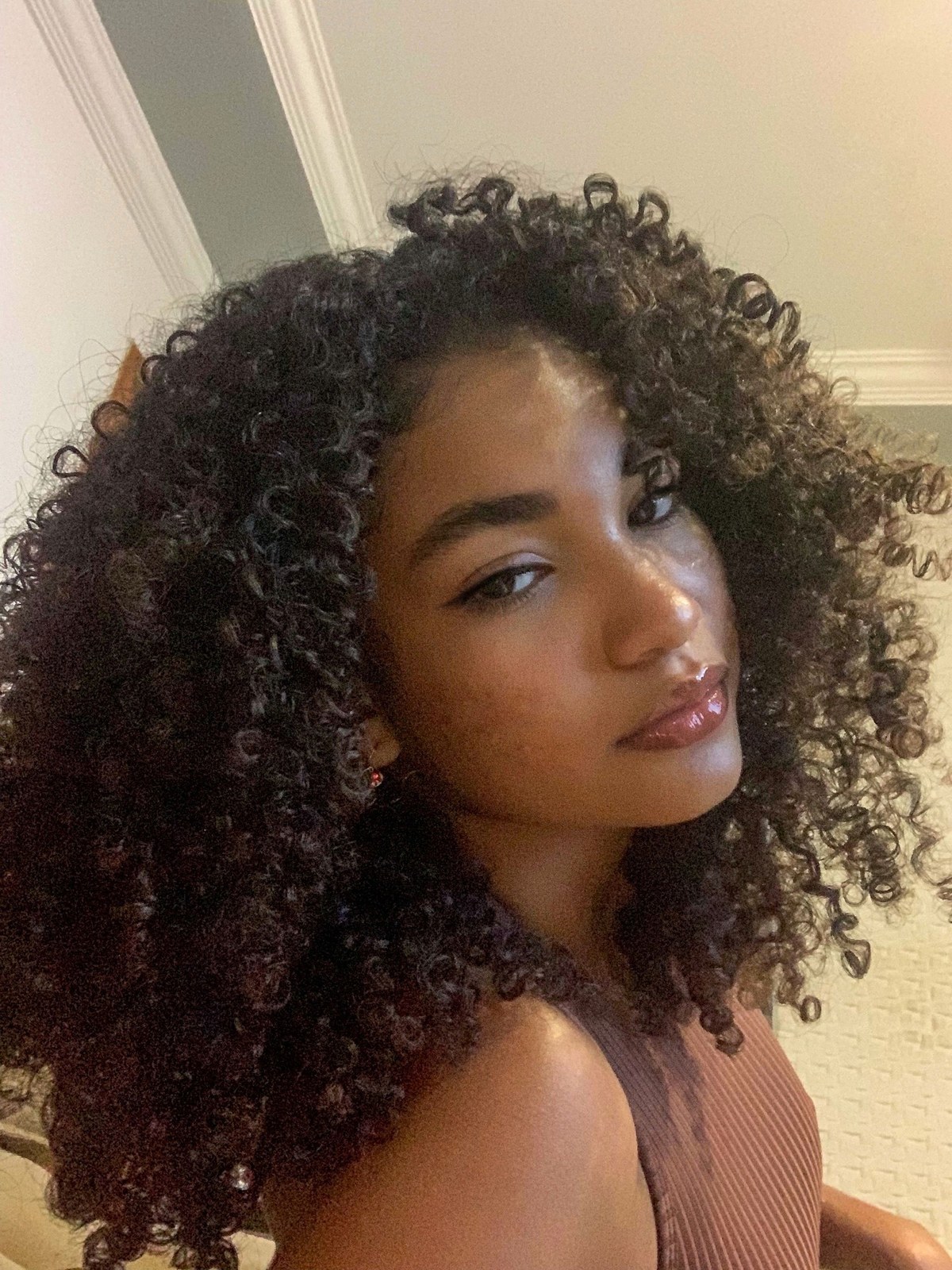 Selfie-style close-up of a woman with big, defined, dark curls looking over her shoulder in a room with white moulding