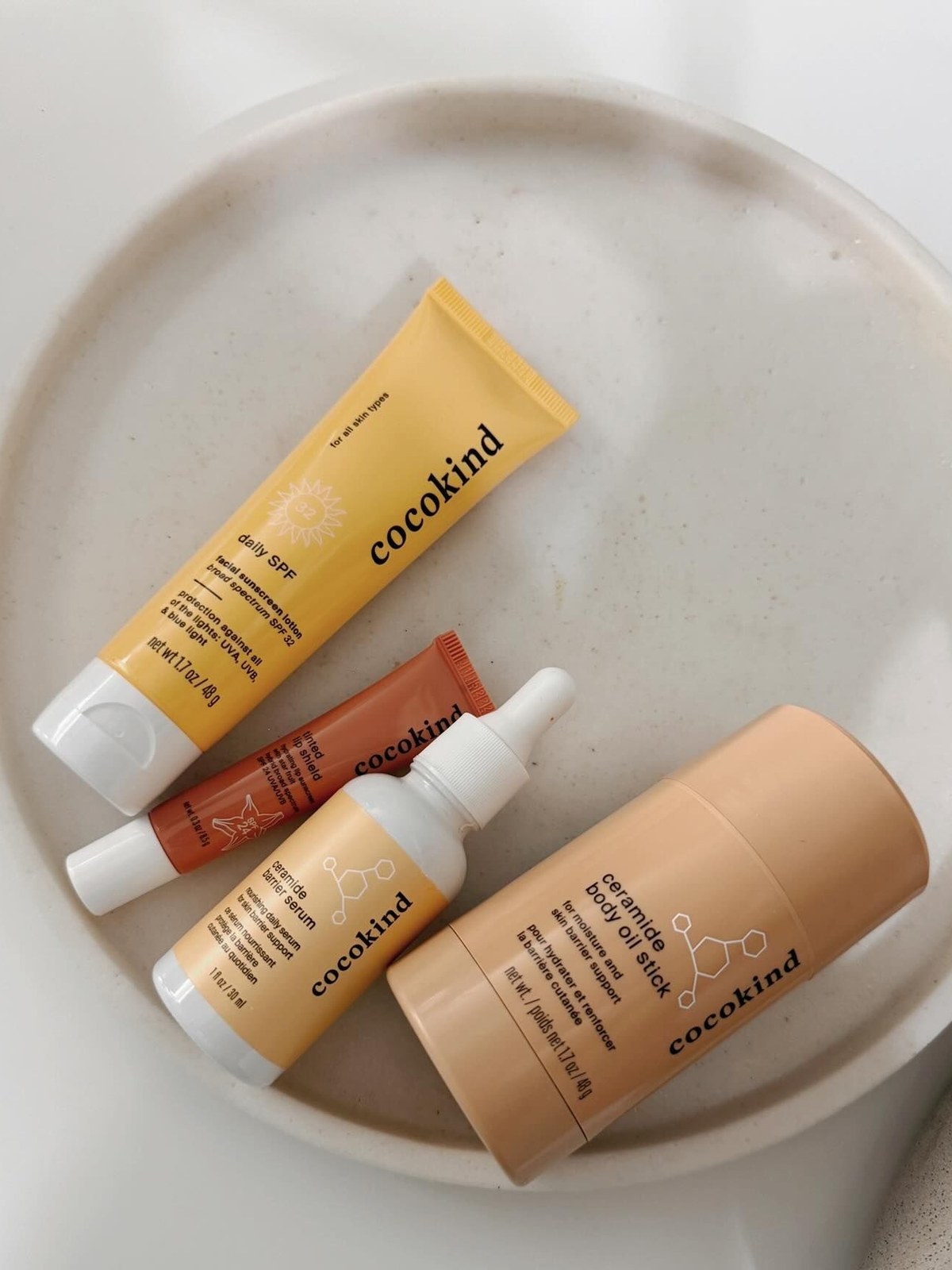 Cocokind daily SPF tube, tinted lip shield, ceramide serum and ceramide body oil stick on a cream ceramic plate