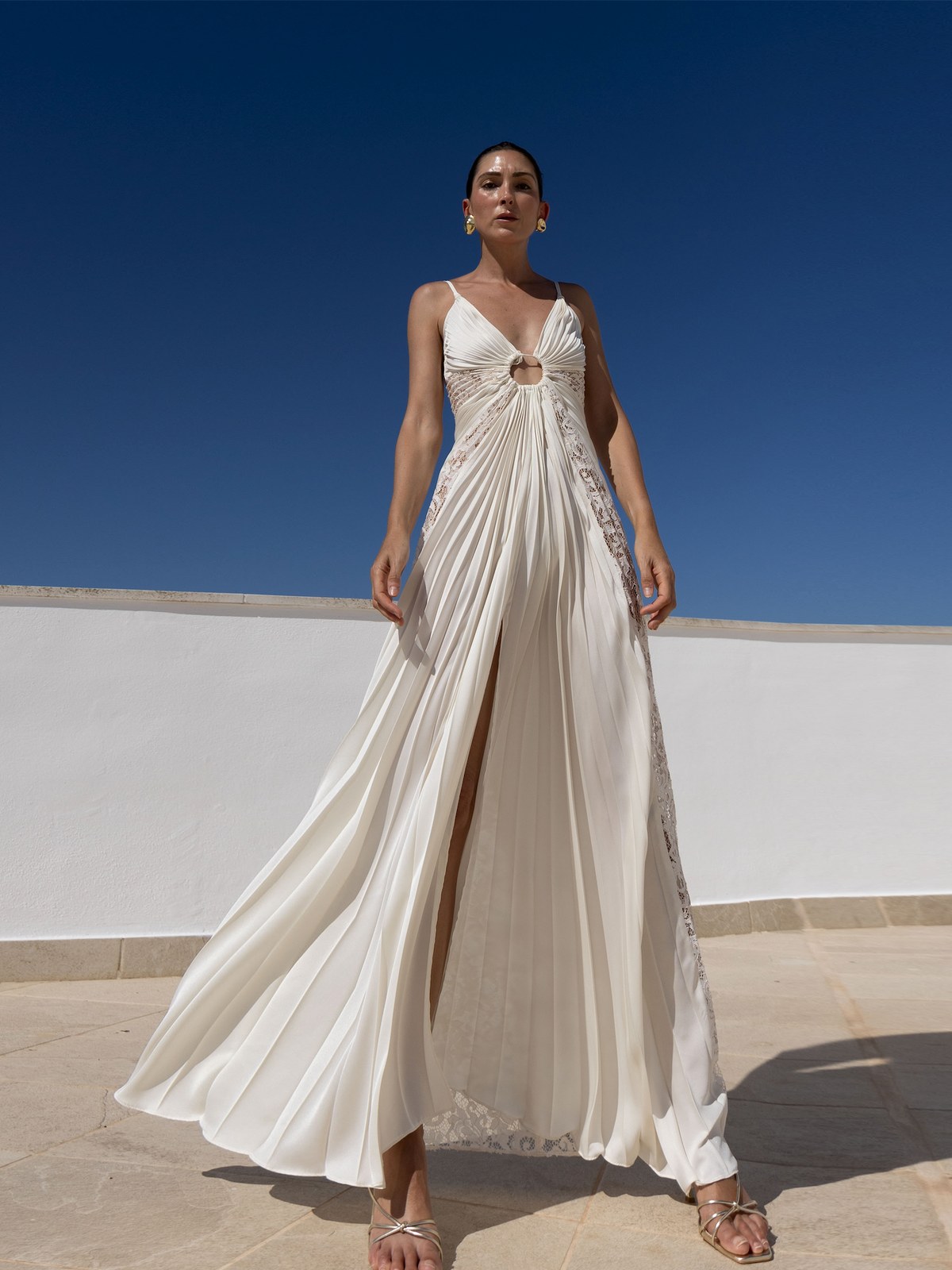 Model in an ivory pleated gown against a blue sky and white wall