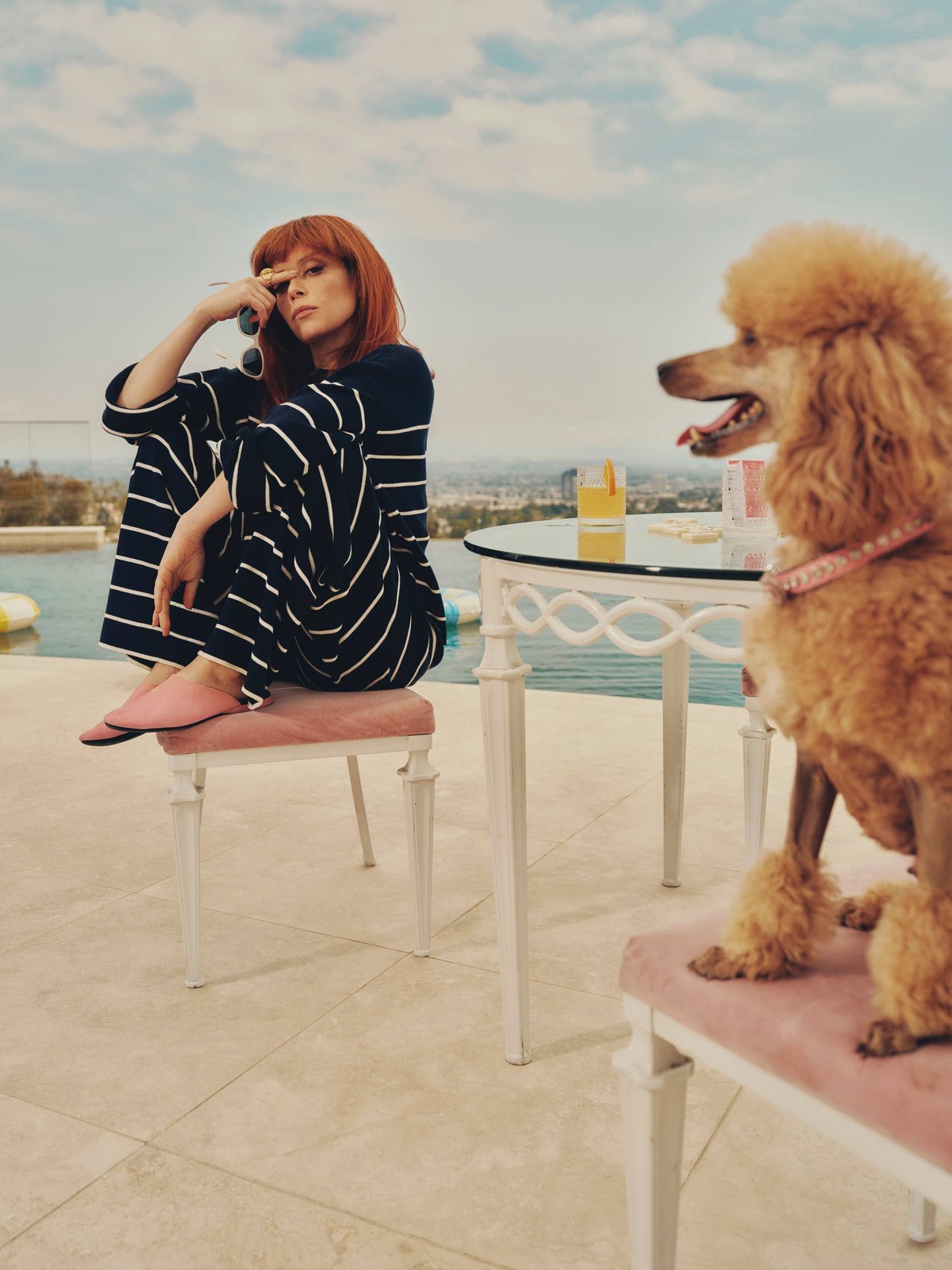 Natasha Lyonne in a navy striped set on a sunny terrace beside a poodle