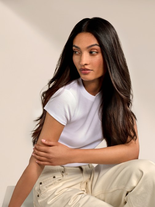 Woman with long dark hair seated in a light studio, wearing a white top and neutral trousers