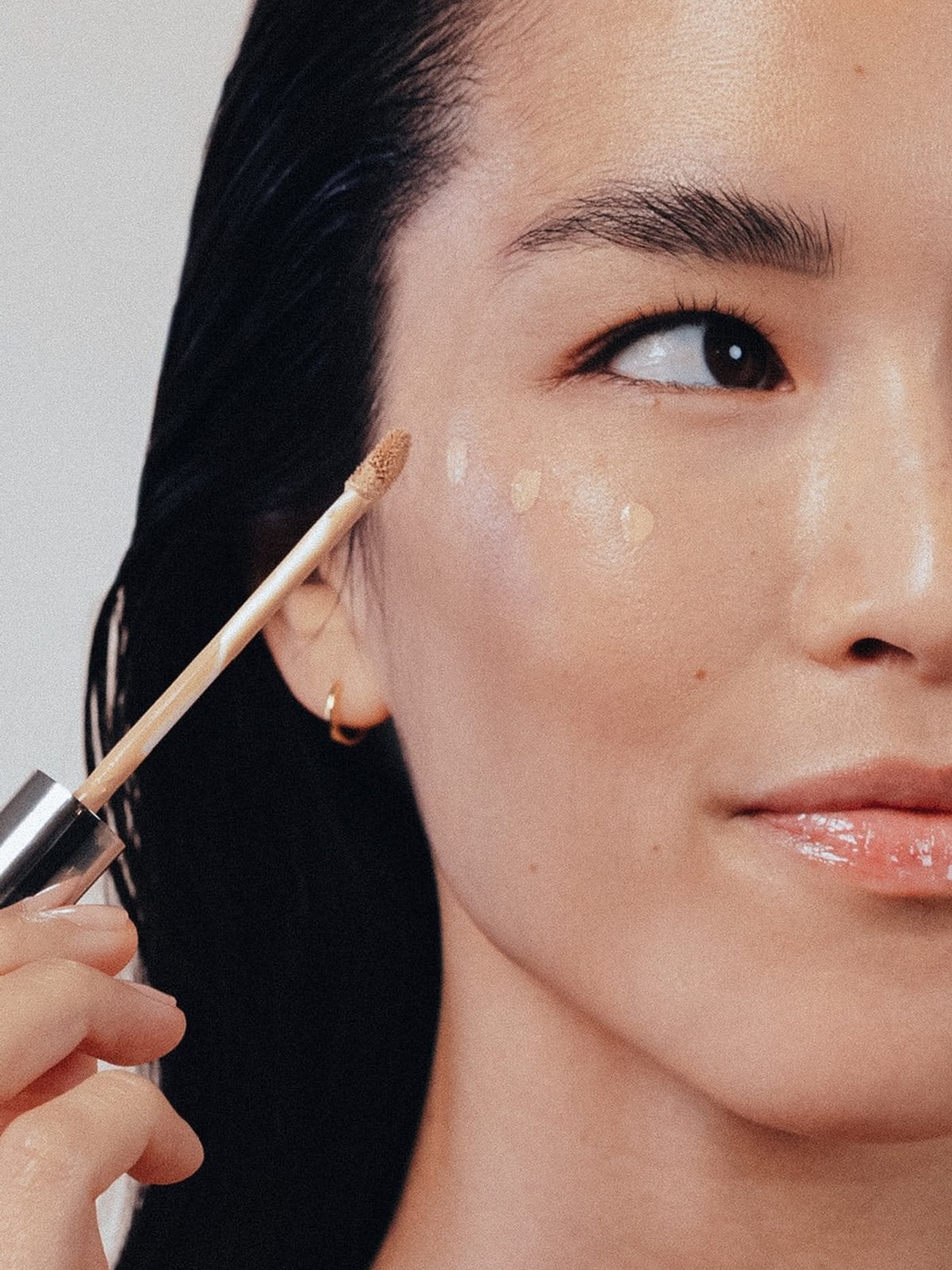 Woman with wet-look dark hair holding a concealer wand beside her cheek, with dots of concealer on her cheekbone