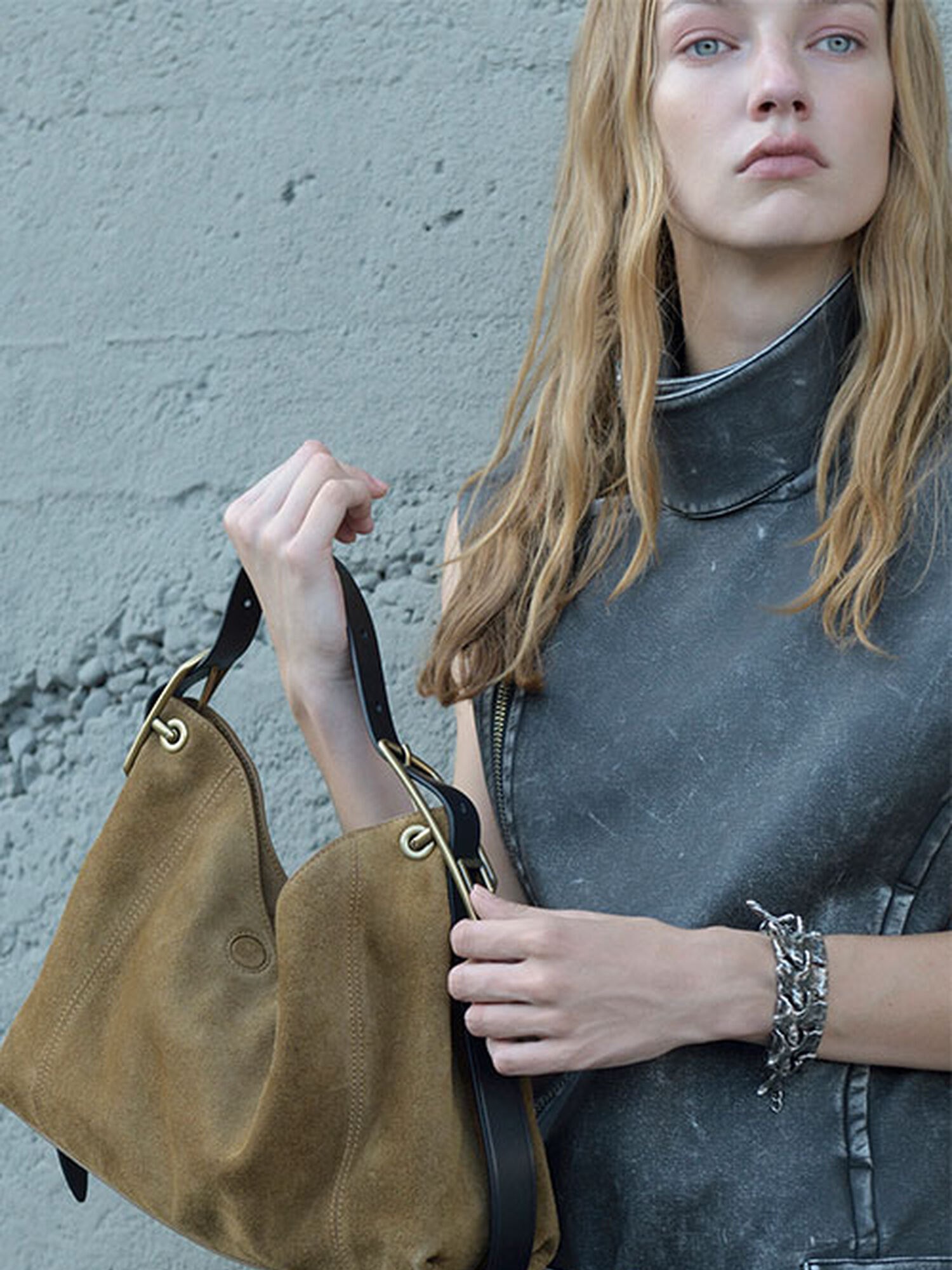 Blonde model in a washed grey top holding a suede hobo bag against a concrete wall