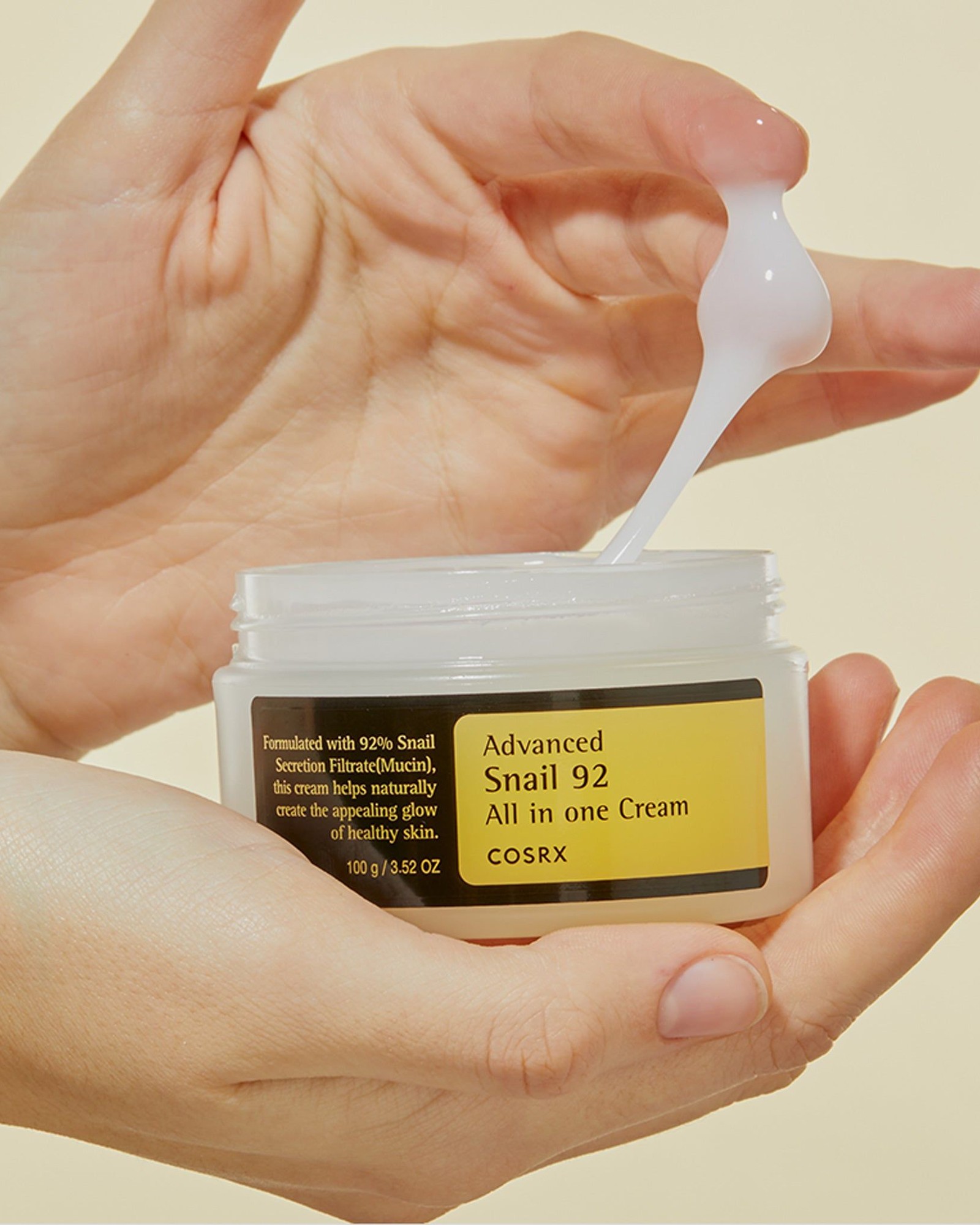 Two hands holding an open jar of COSRX Advanced Snail 92 All in one Cream while a finger lifts a stretchy ribbon of white gel cream
