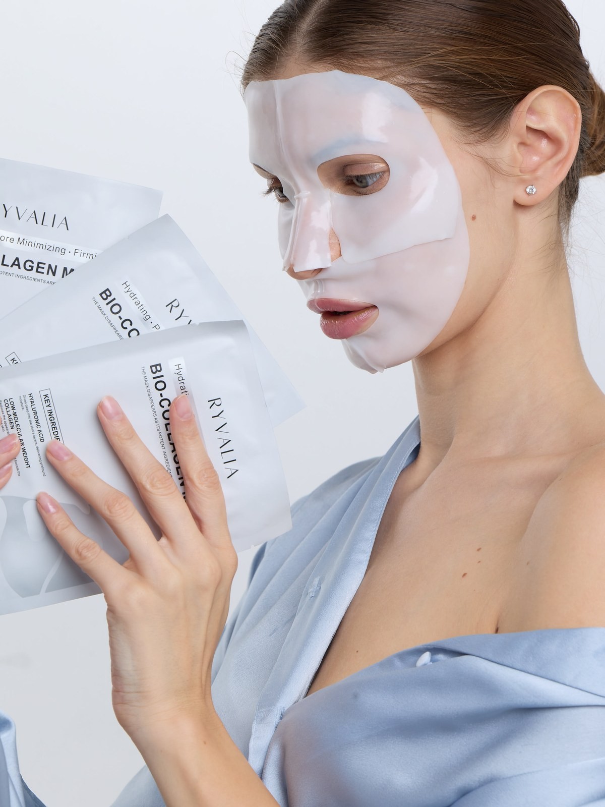 Woman in a sheet mask holding a fan of skincare mask packets