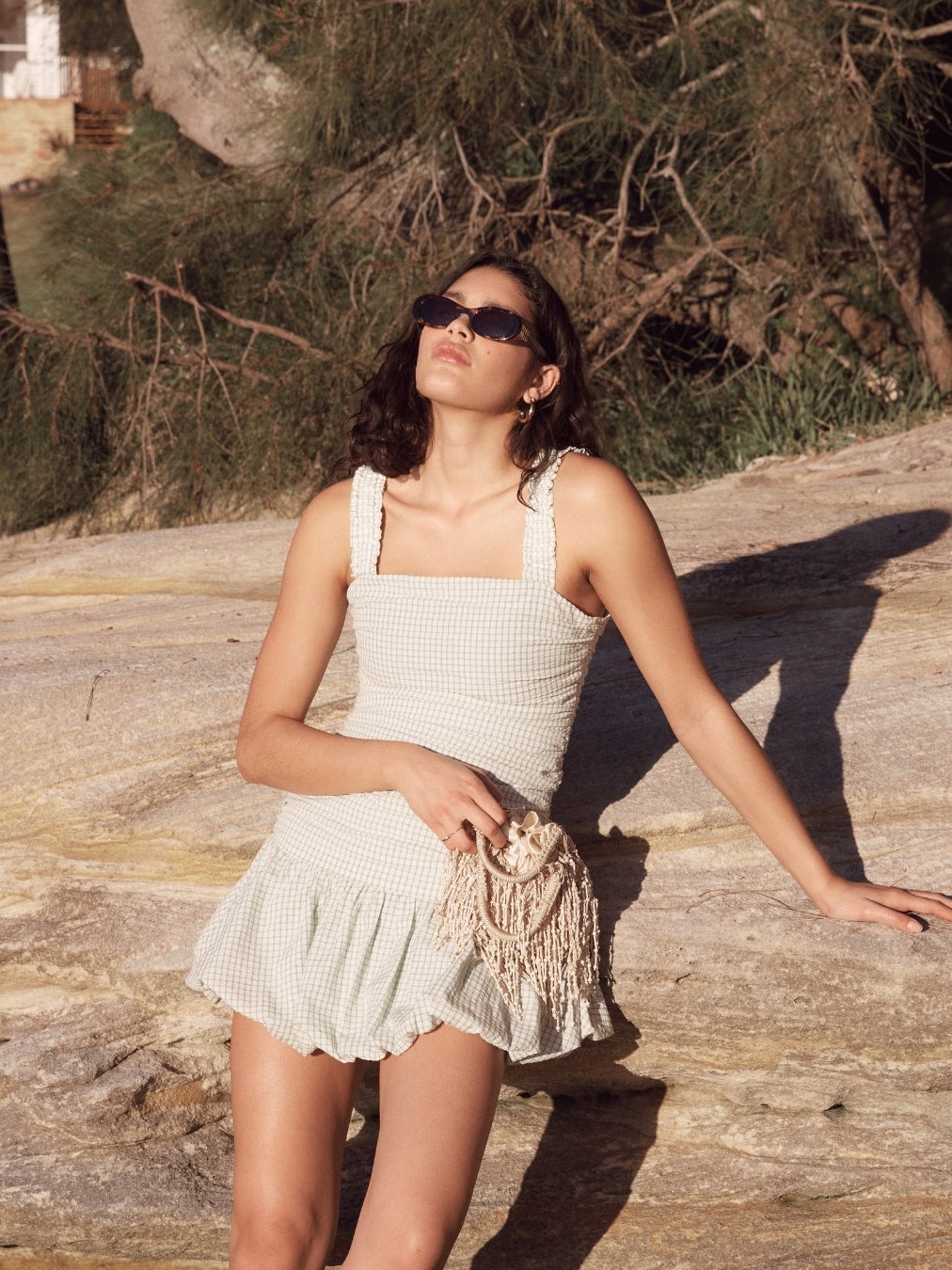 Model in a white MINKPINK summer outfit seated in a sunlit natural setting