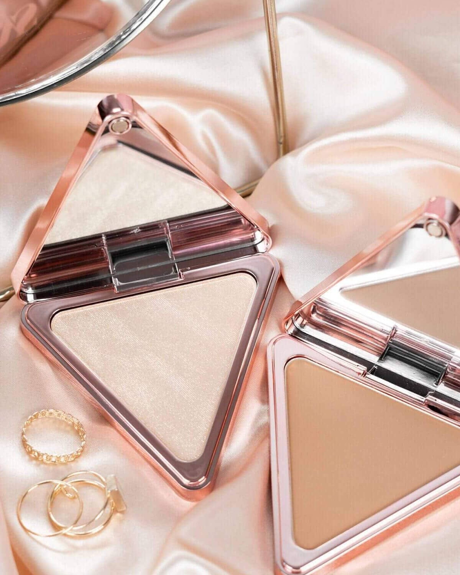 Two triangular rose-gold compacts, one holding a pale shimmery highlighter and one a warm bronzer, open on peach satin beside gold rings.