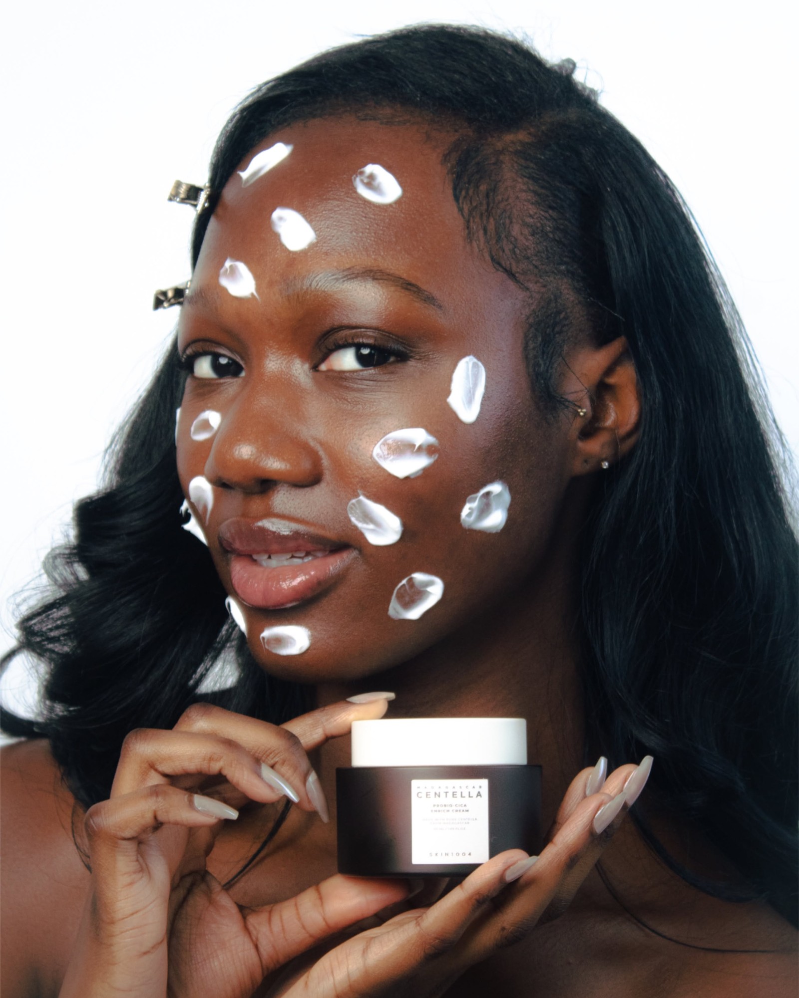 Woman with dark shoulder-length hair and dabs of white cream across her cheek and forehead, holding a jar of SKIN1004 Probio-Cica Enrich Cream