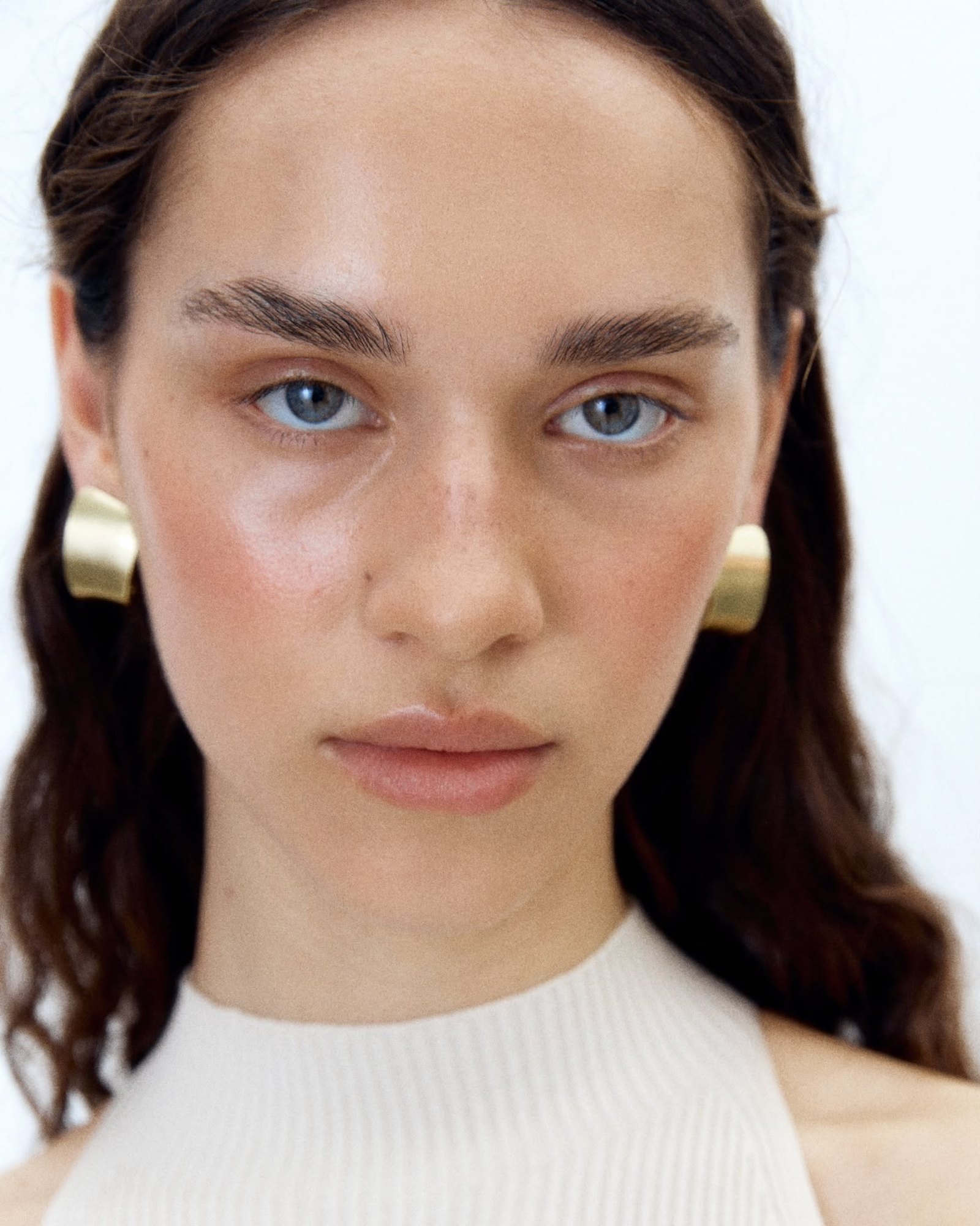 Model with wavy brown hair, blue eyes and a dewy complexion with a soft blush, wearing sculptural gold earrings and a cream halter top