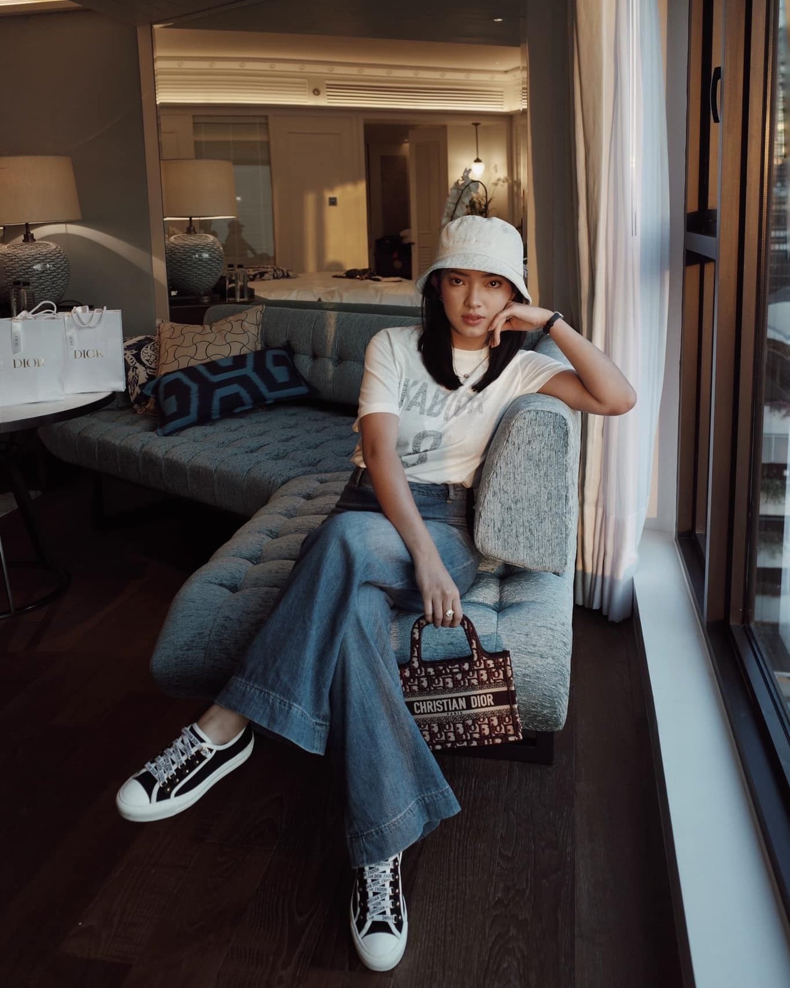 Woman in a hotel room in a bucket hat, white T-shirt and wide jeans with Walk'n'Dior sneakers and a Dior bag