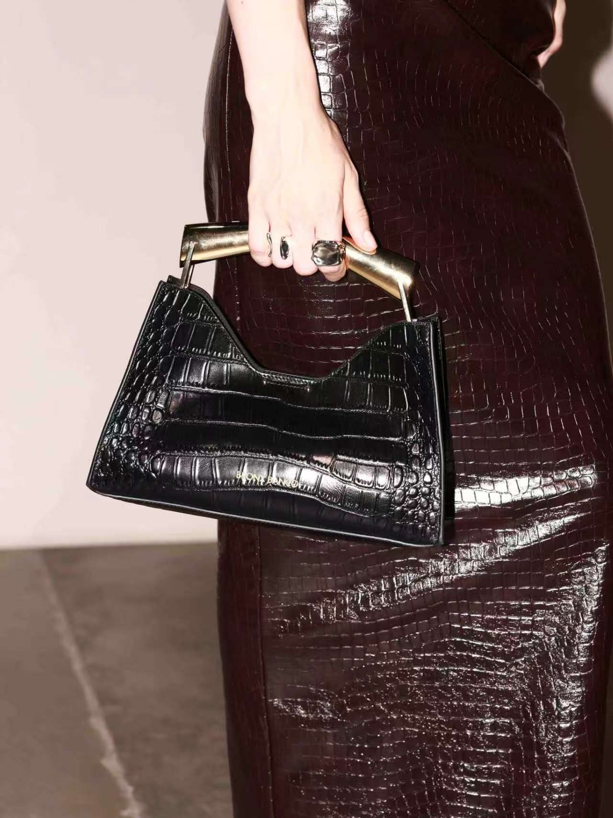 Close up of a model holding a black croc embossed handbag against a burgundy croc skirt