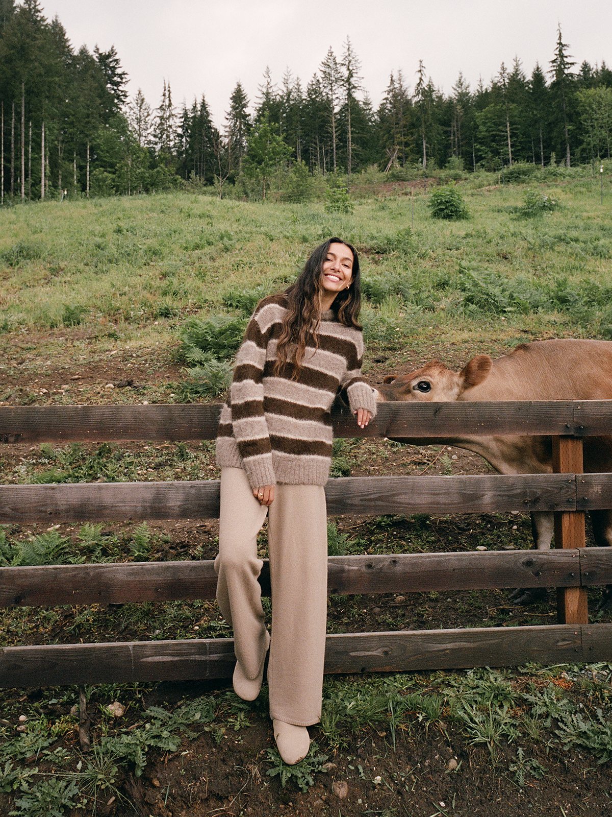 Model in a striped brown and cream sweater leaning on a pasture fence beside a cow