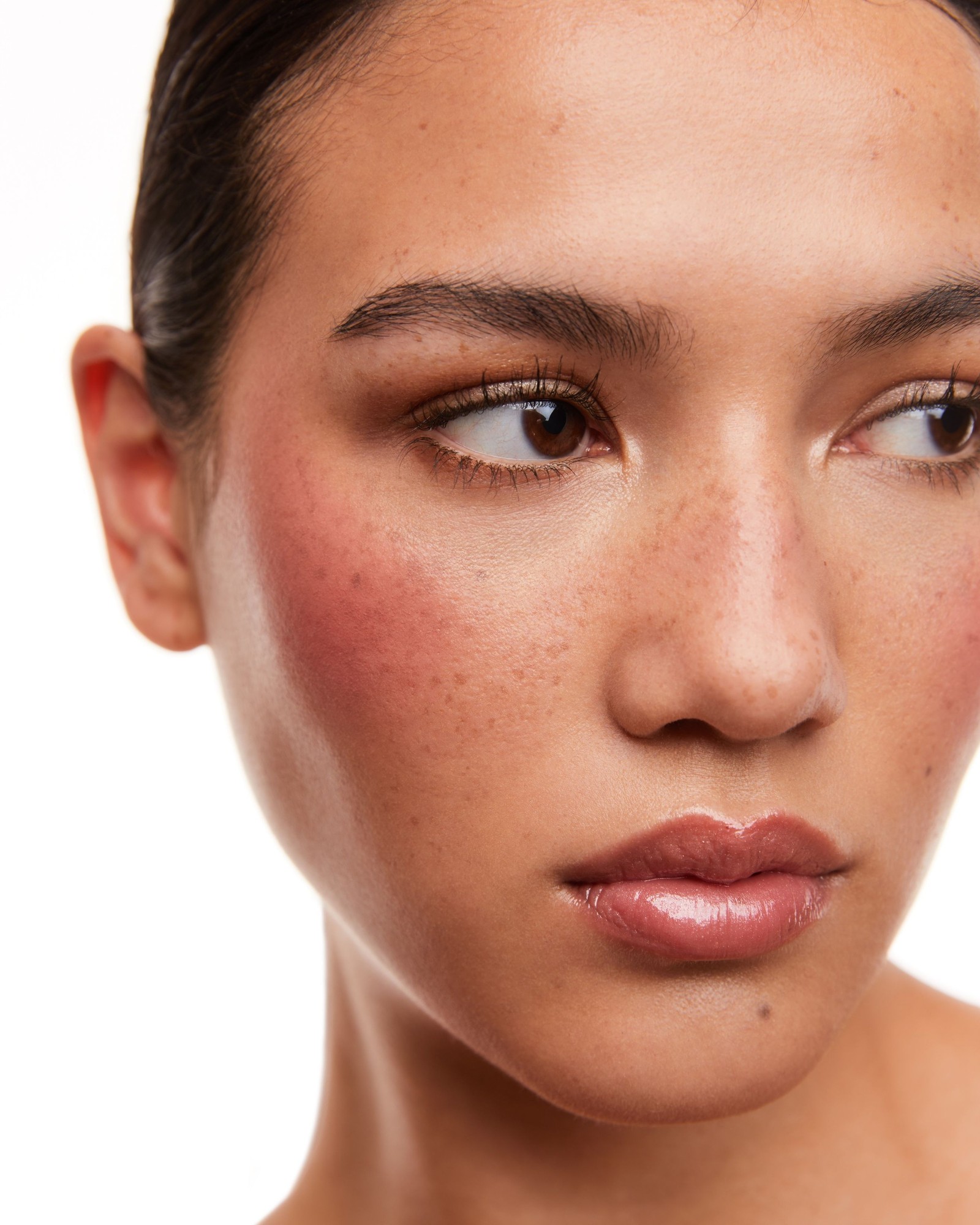 Tight close-up of a model with freckles, a rosy cheek flush and a glossy pink lip, looking to the side against a white background