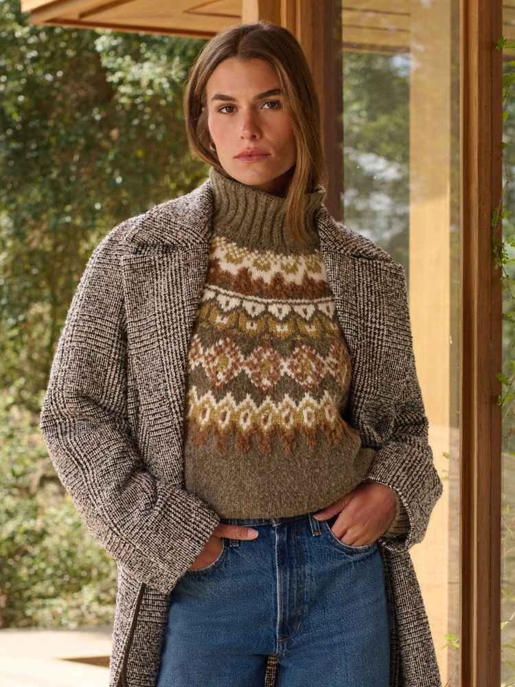 Woman wearing a textured sweater, coat and jeans by a glass doorway
