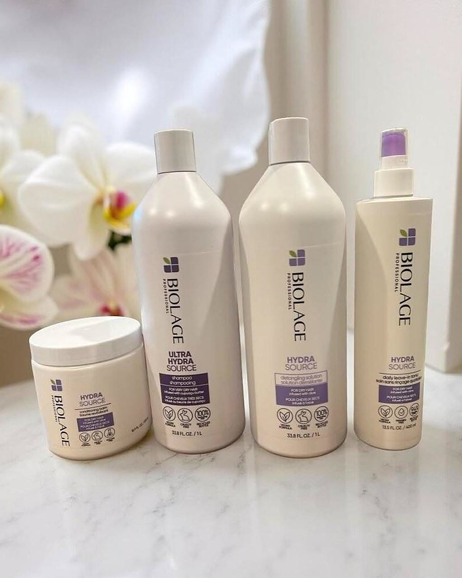 Four cream and purple Hydra Source hair care bottles and a jar lined up on a marble counter in front of white orchids