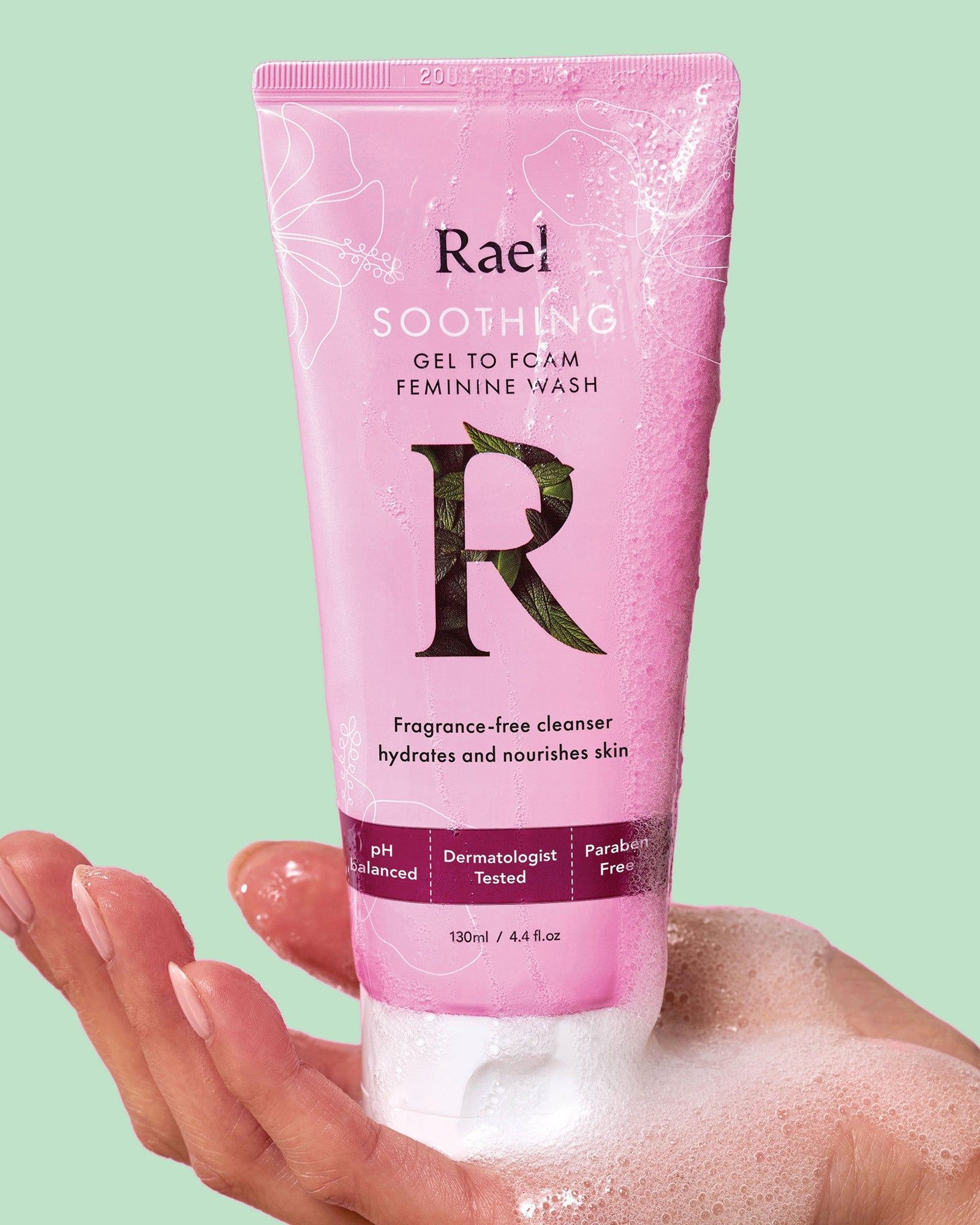 A hand holding a pink tube of Rael Soothing Gel to Foam Feminine Wash covered in foam against a mint green background