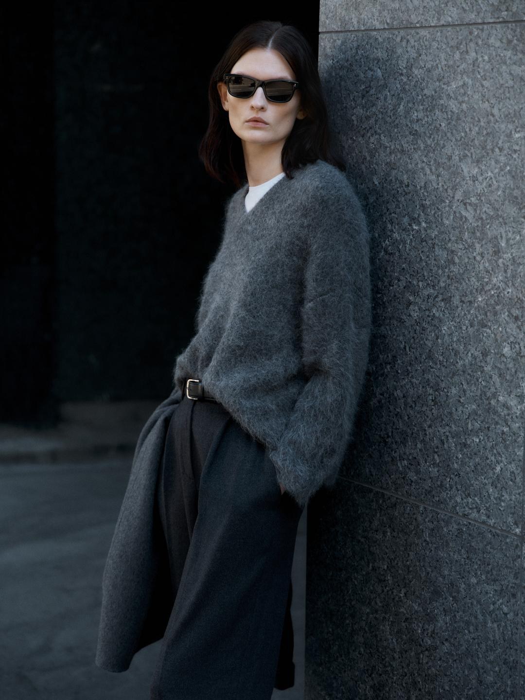 Model in an oversized grey knit and dark trousers standing by a city wall