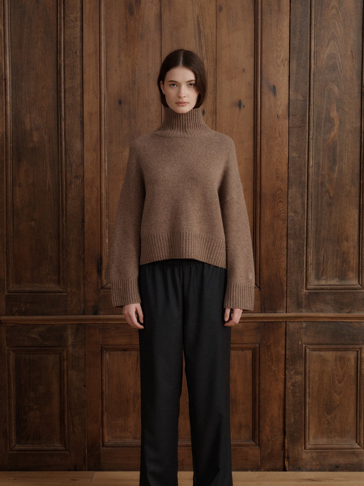 Model wearing a toasted walnut turtleneck and black trousers in a wood-panel studio
