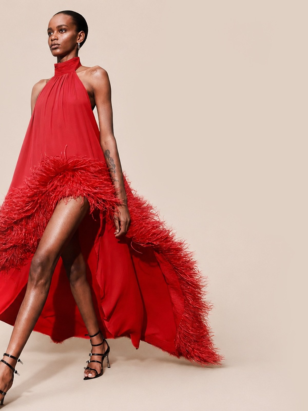 Model in a red halter ruffled gown with feather trim mid stride on a blush backdrop