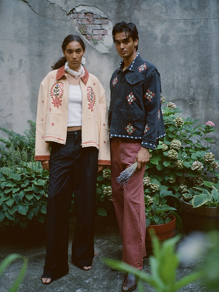 Two models in embroidered jackets against a garden wall