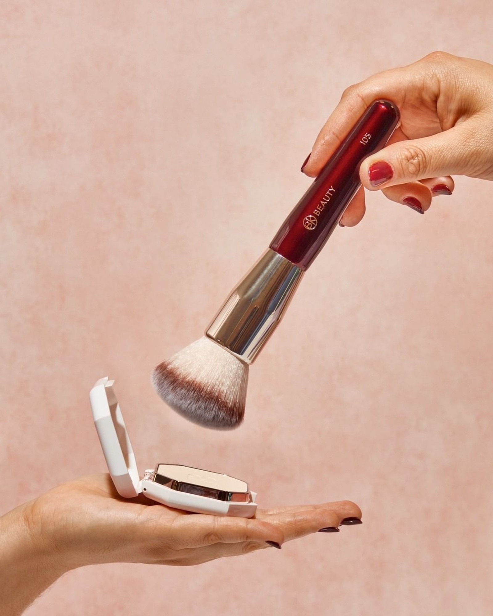 Hand holding a burgundy BK Beauty brush numbered 105 above an open white powder compact held in another hand, on a pink background