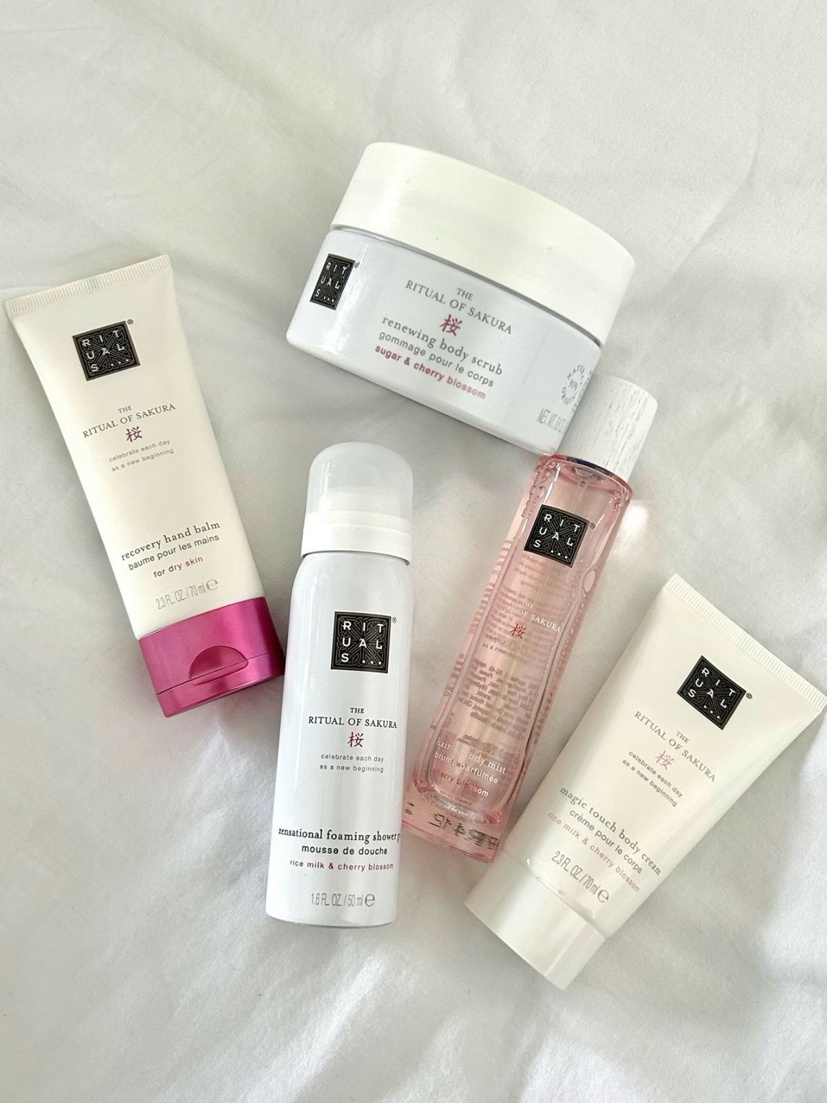 Flat lay on white sheets of The Ritual of Sakura hand balm, body scrub, foaming shower gel, hair and body mist and body cream