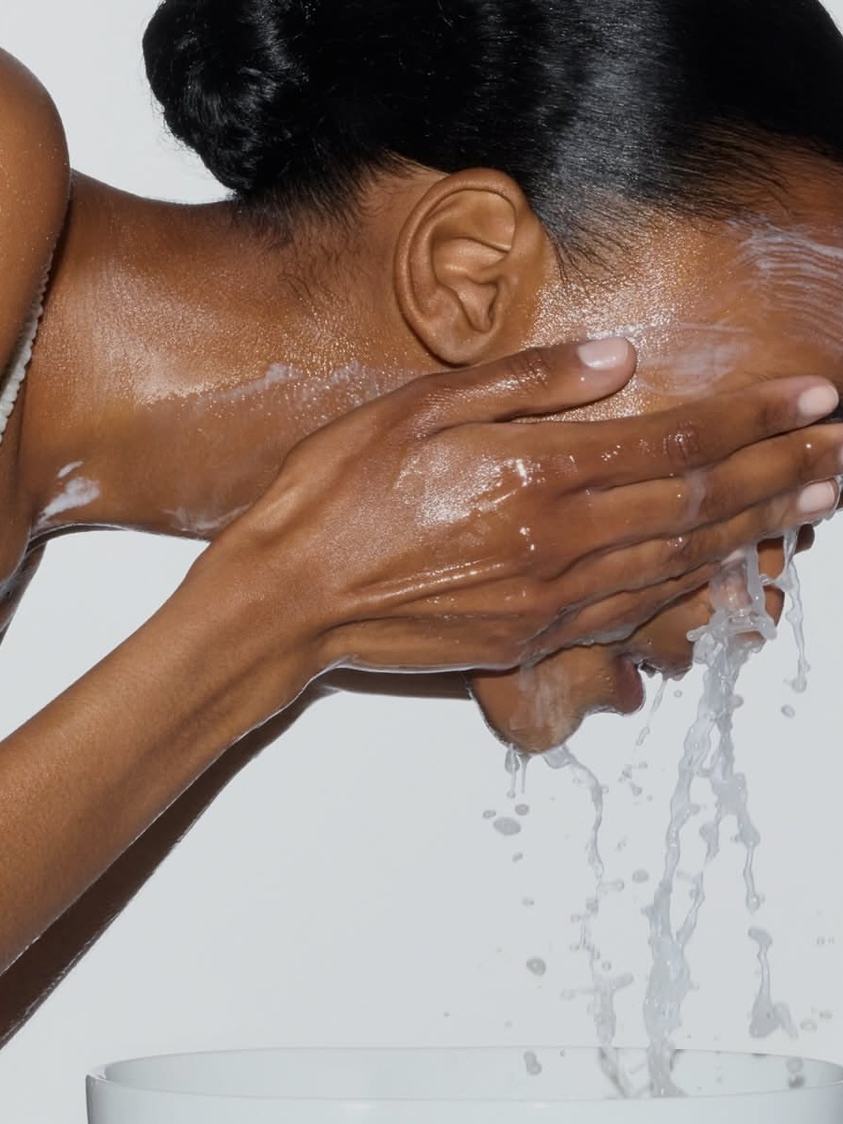 Woman leaning forward and splashing water on her face over a white basin, with soapy lather on her neck