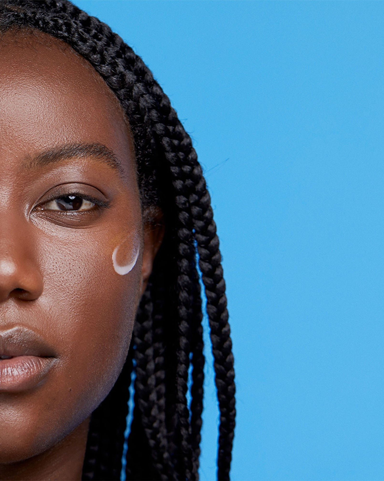 Half portrait of a woman with long box braids and a small swipe of white cream on her cheek against a light blue background