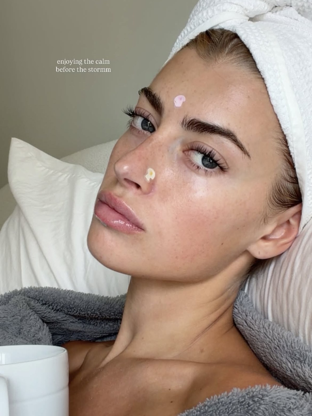 Woman with a white towel wrapped around her hair, a pink dot of treatment on her forehead and a cream dab on her nose, holding a mug in a grey robe