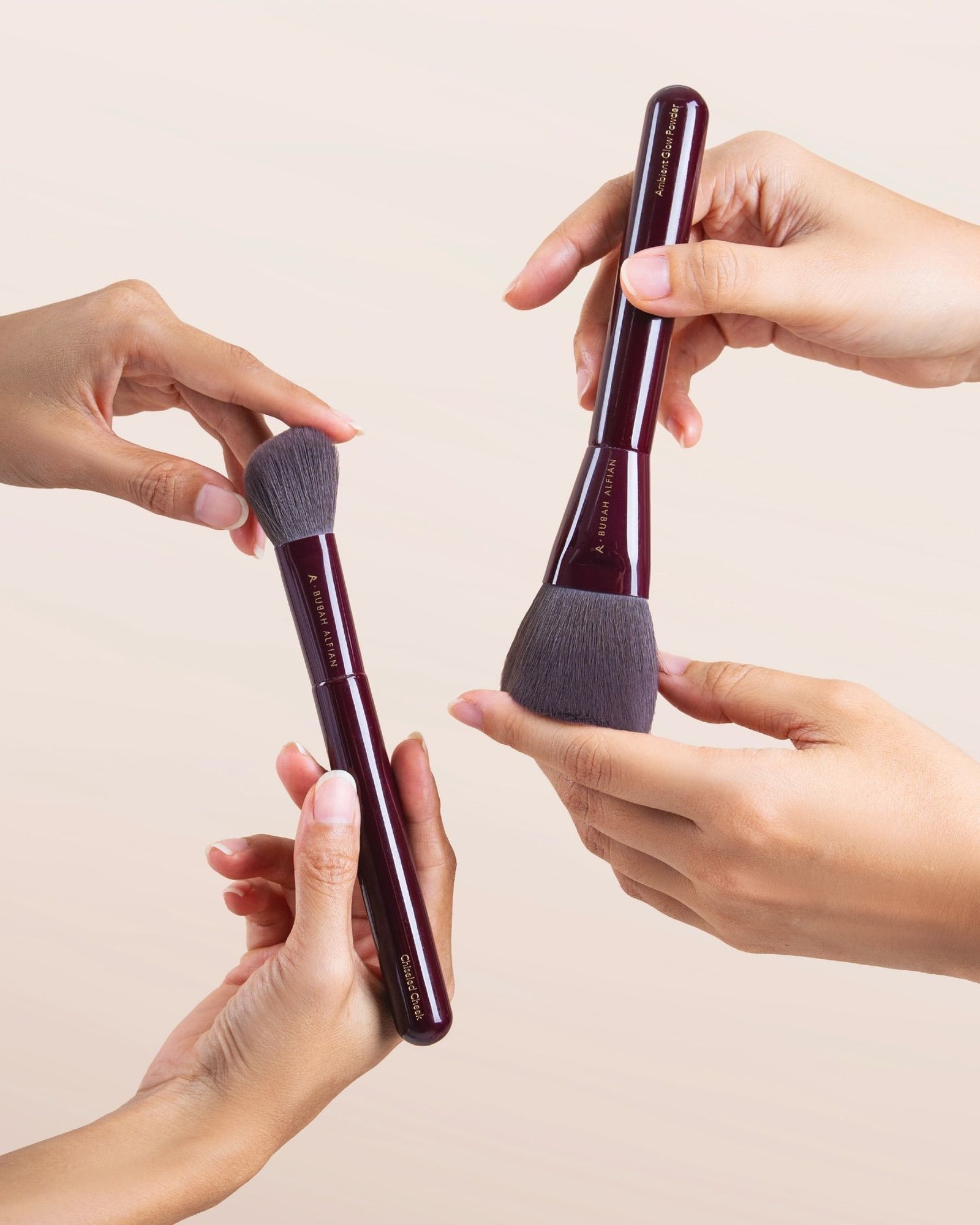 Three hands holding two plum-handled makeup brushes, a small angled cheek brush and a large powder brush, against a soft cream background