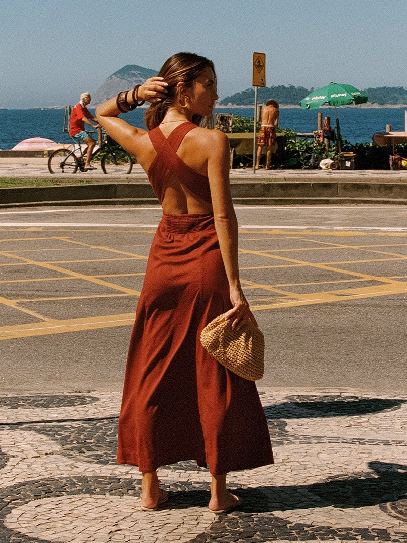 Model in a rust-colored dress carrying a woven bag along a waterfront
