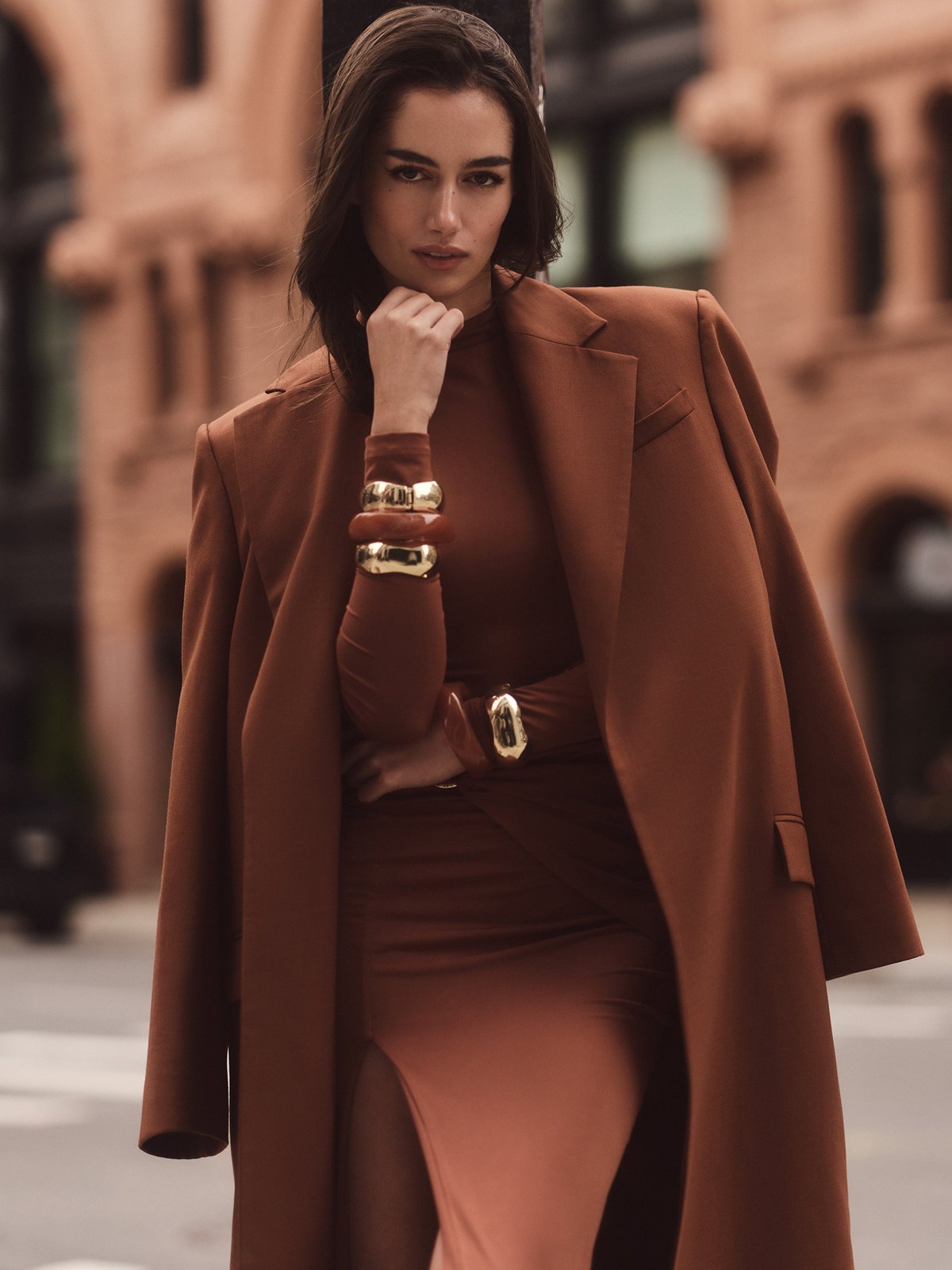 Nookie model in a brown coat over a fitted brown dress on a city street.