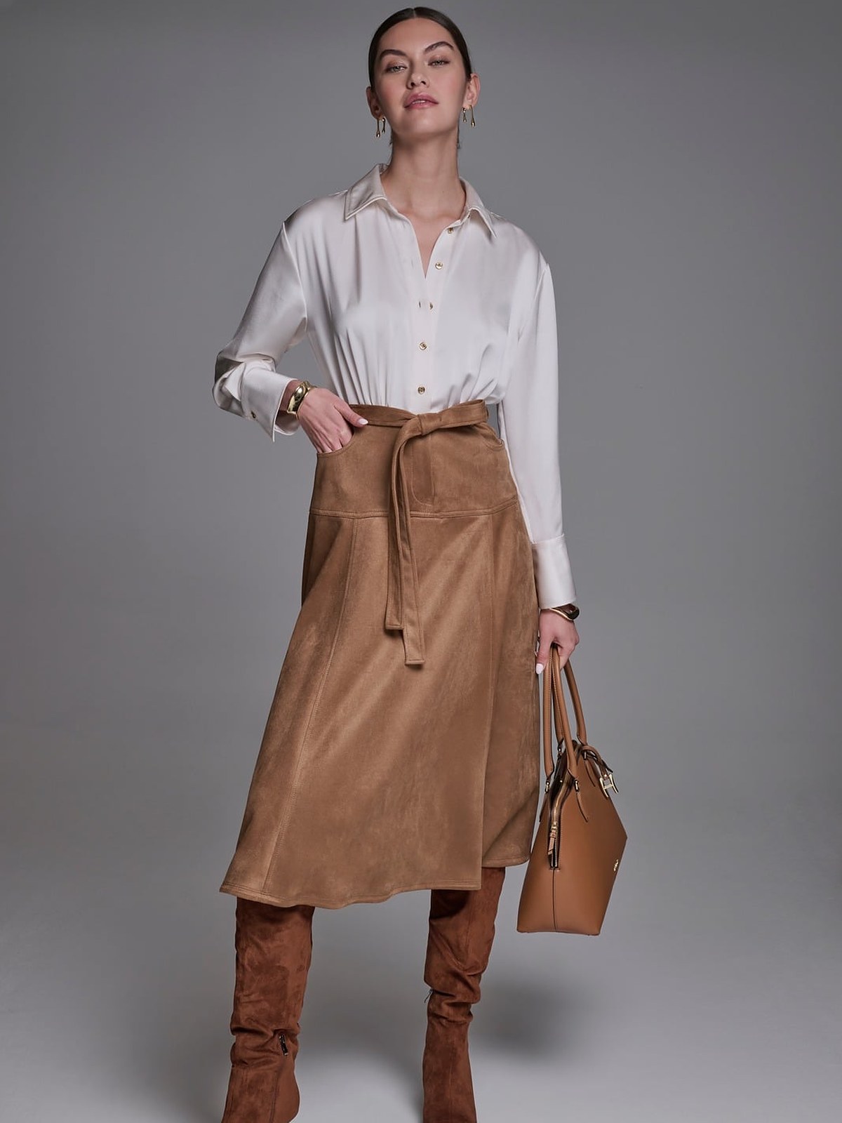 Model in a white satin shirt, camel suede wrap skirt and suede knee boots with a tan tote