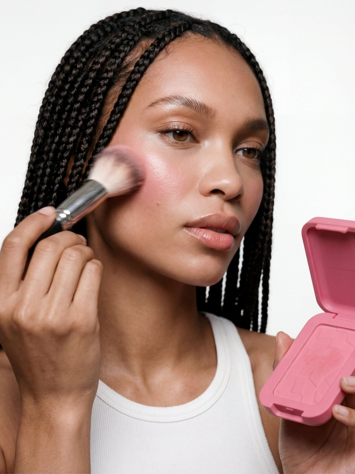 Woman with long box braids brushing pink blush on her cheek while holding a pink compact
