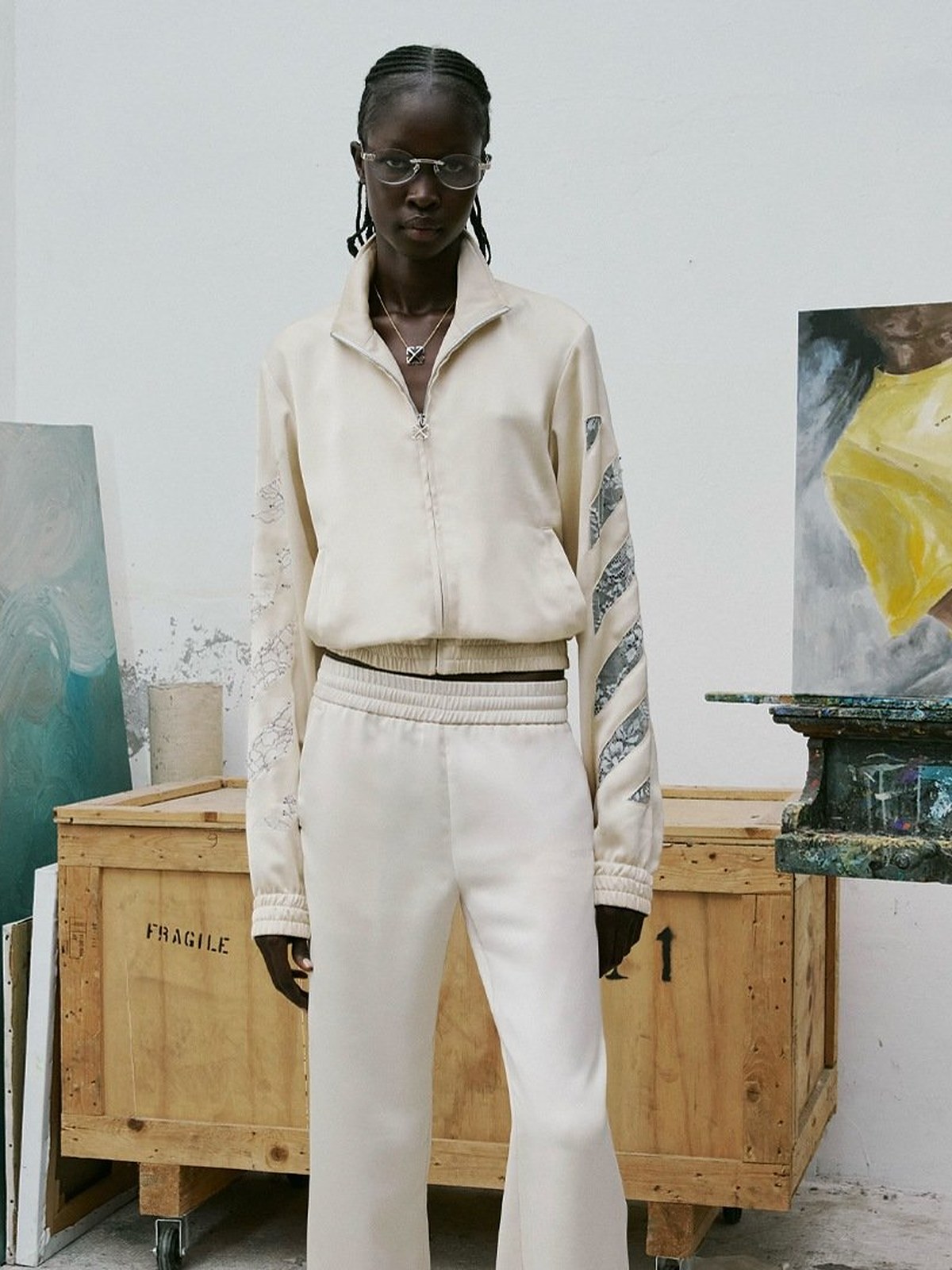 Model in a cream zip track jacket and white track pants in an art studio