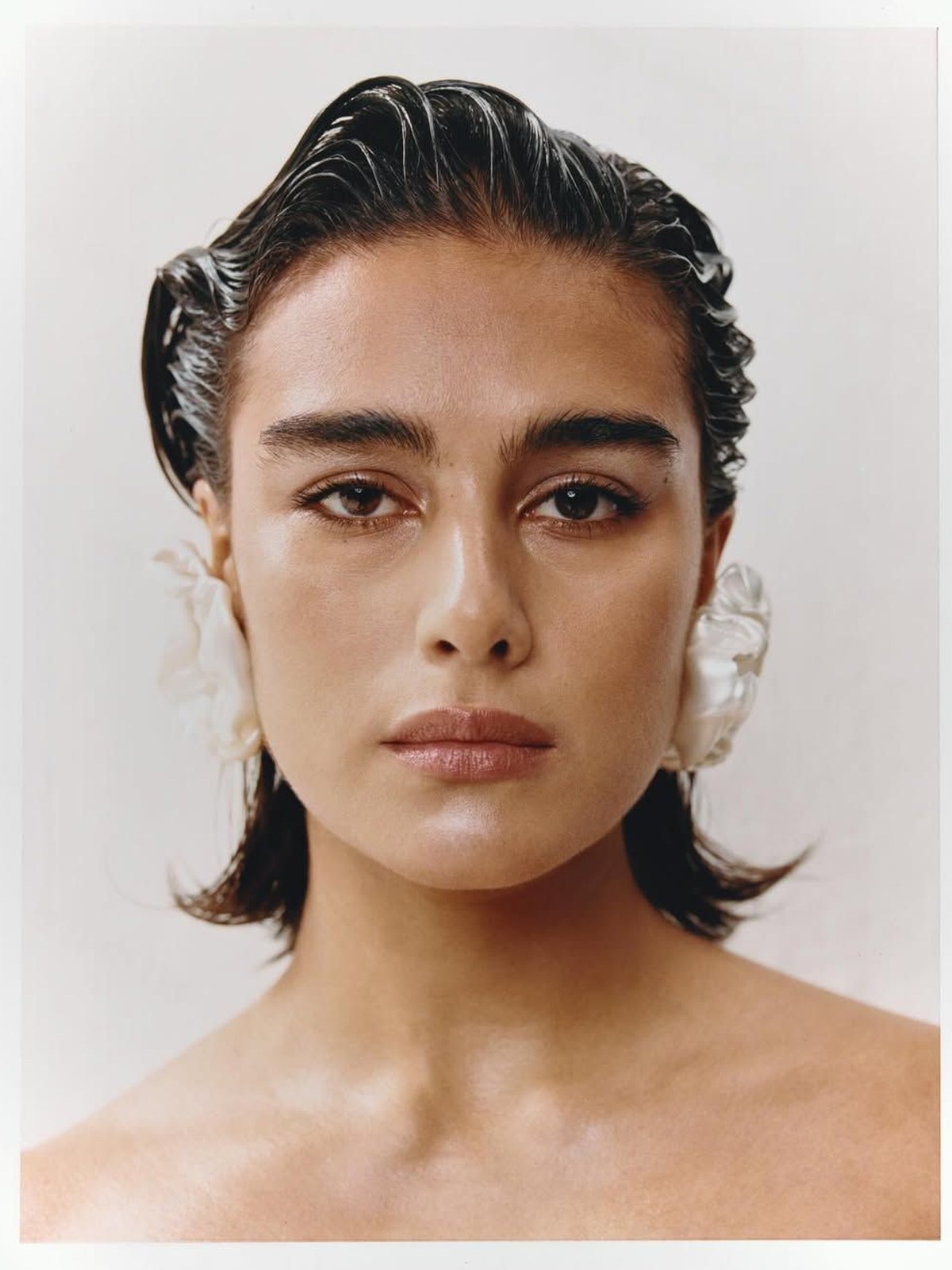 Back view of a woman with wet-look slicked-back dark hair in a low bun, wearing large gold dome earrings and a black knit collar