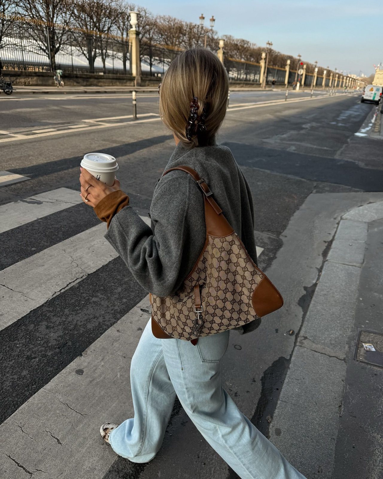 Woman crossing a street in a grey jacket and light jeans with a Gucci GG canvas Jackie bag, holding a coffee