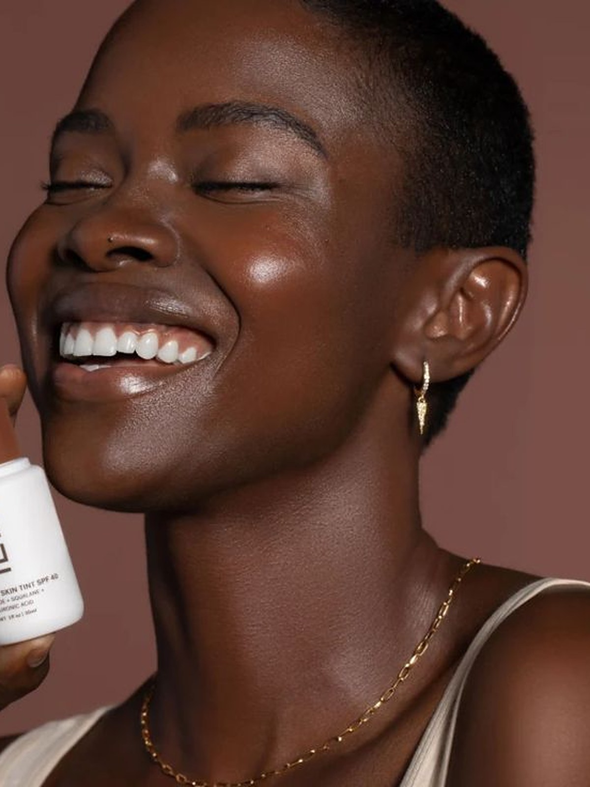 Smiling model holding an ILIA serum bottle against a warm brown backdrop