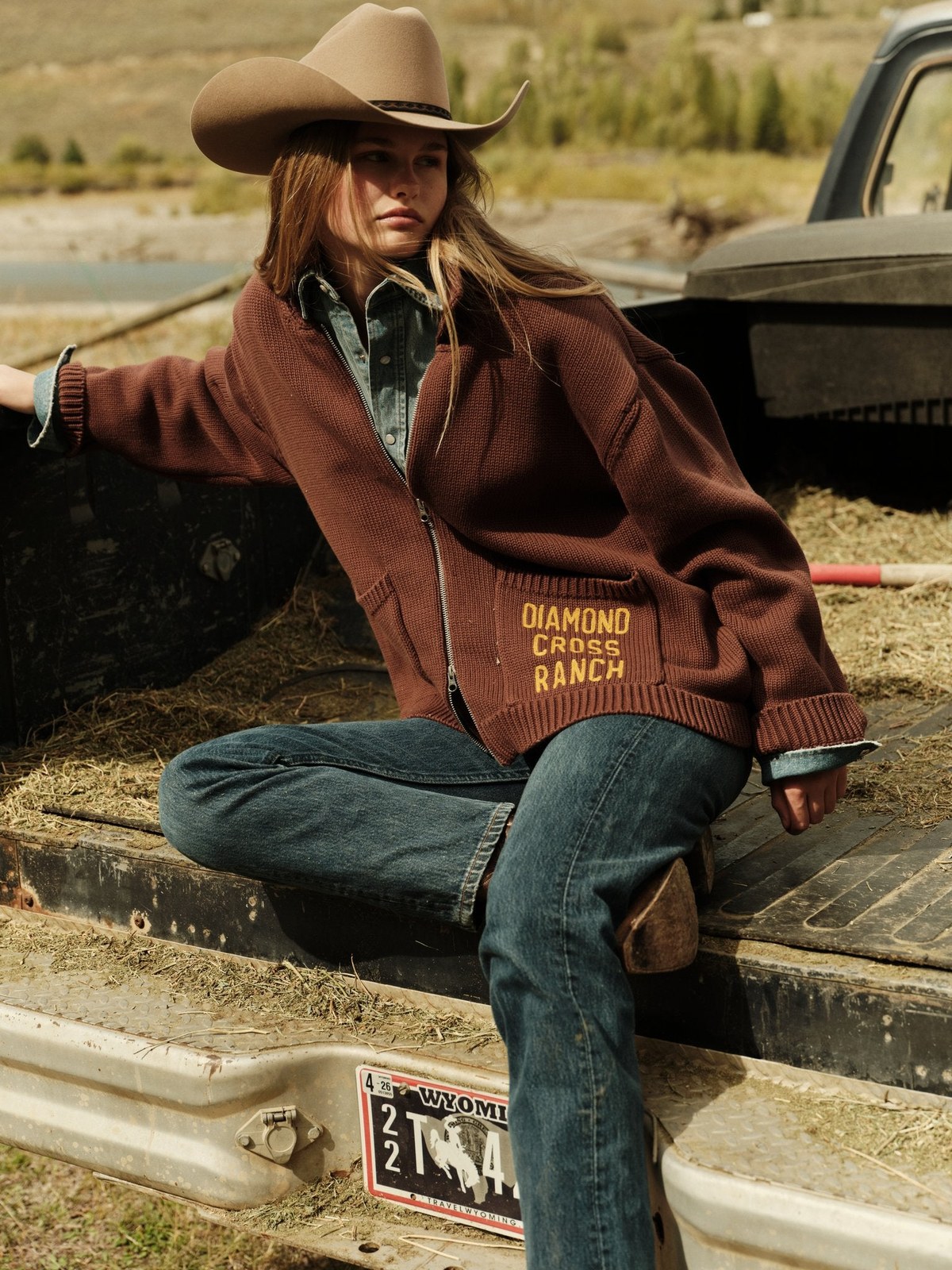 Model in a brown ranch-logo jacket and cowboy hat seated beside a truck.