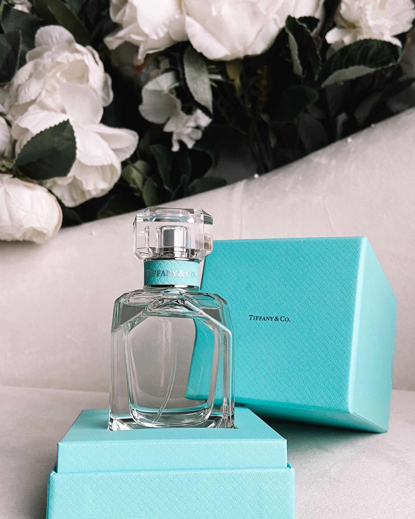Faceted Tiffany & Co. perfume bottle standing in an open blue gift box with white peonies behind it