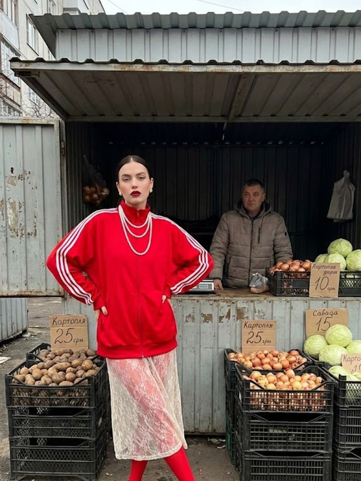 Model in a red sweater, pearls and a pale skirt with red tights at a fruit stall