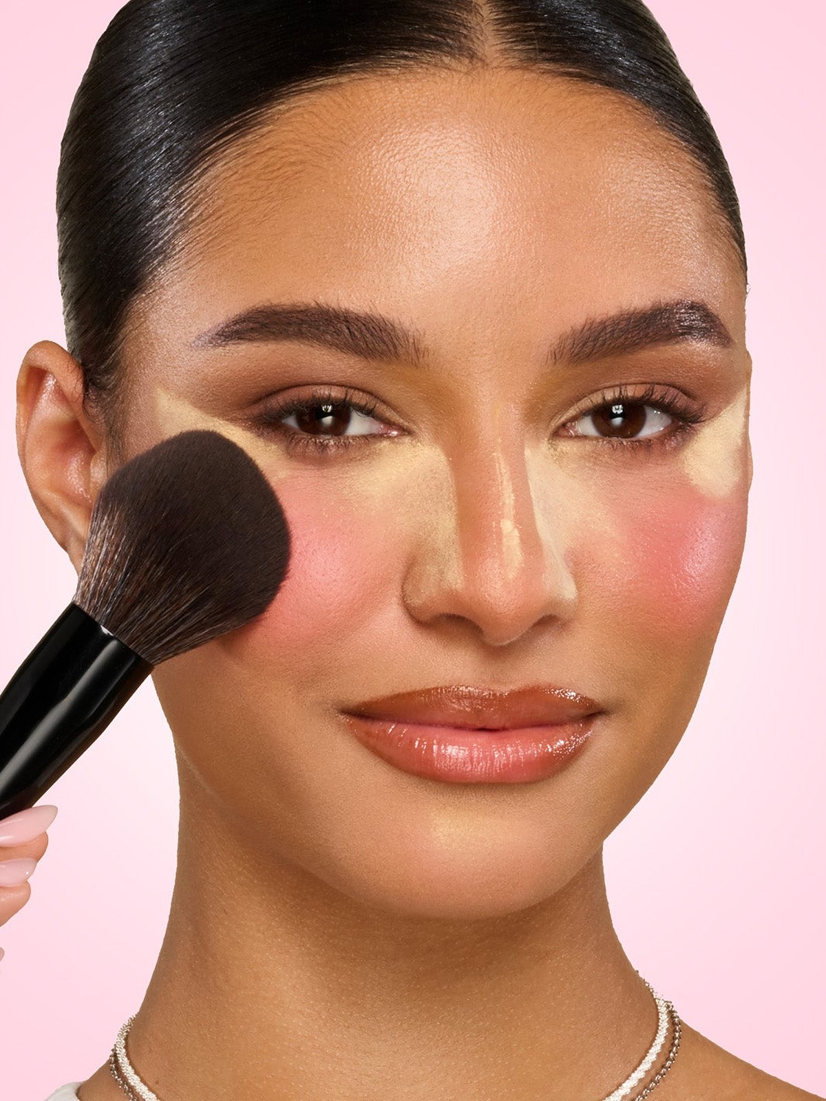 Model with a blush brush on her cheek and glowing makeup on a pink backdrop
