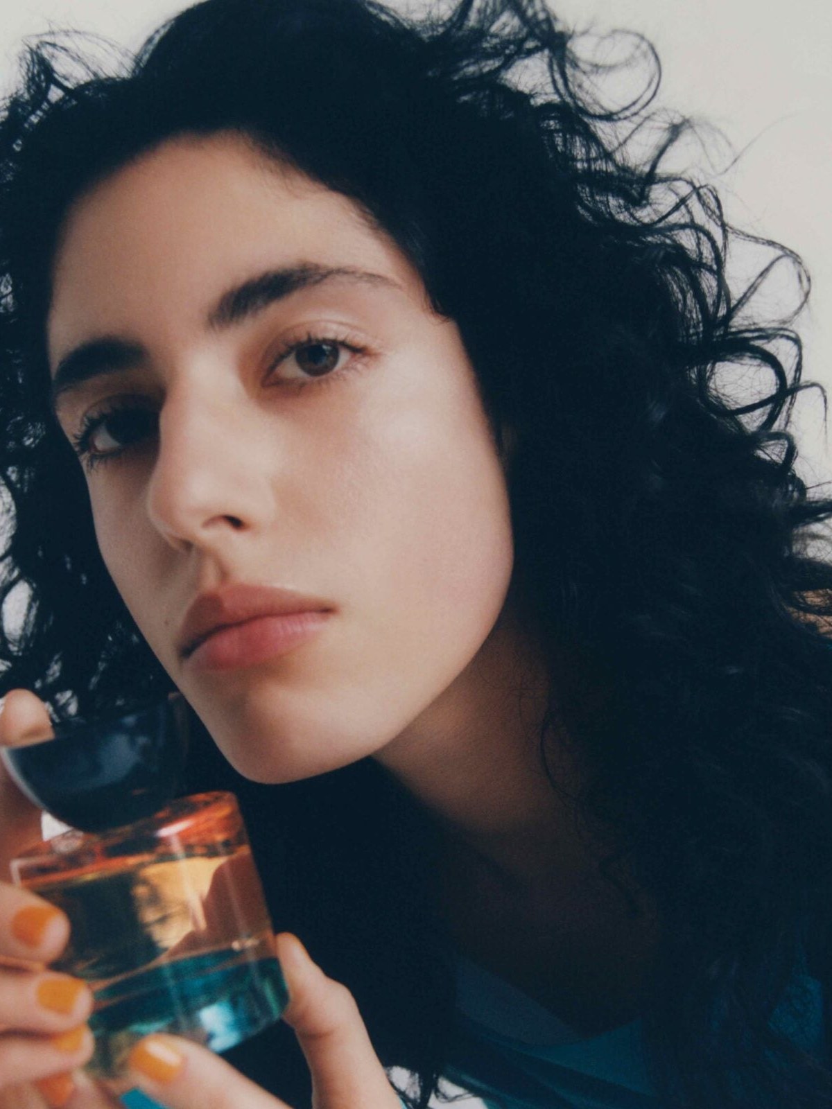 Model with dark curls holding a fragrance bottle in a teal shirt