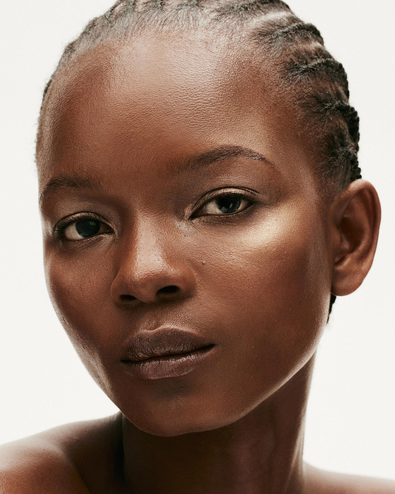 Close-up beauty portrait of a woman with cornrow braids and deep skin, a soft golden glow on her cheekbones, looking at the camera against a white background.