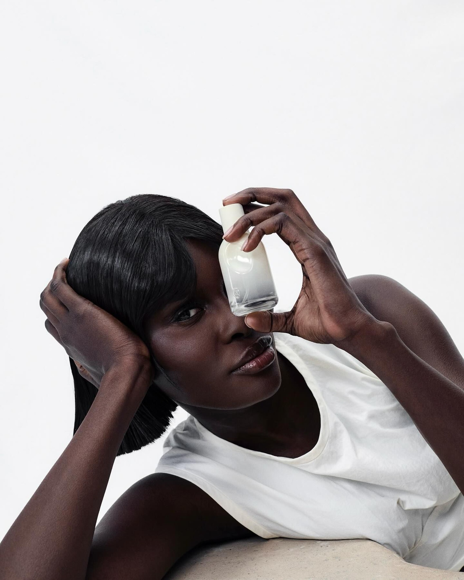 Model with a sleek black bob and a white tank resting her head on one hand while holding a small frosted perfume-style bottle in front of one eye