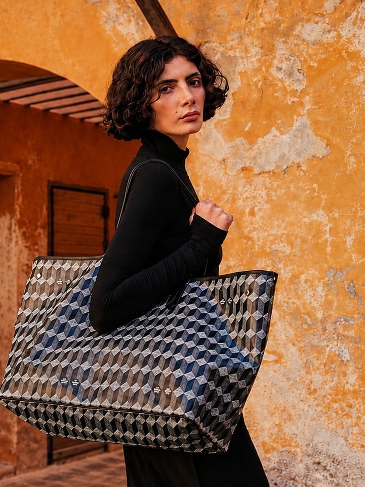 Model with a monogram tote against an ochre arched wall