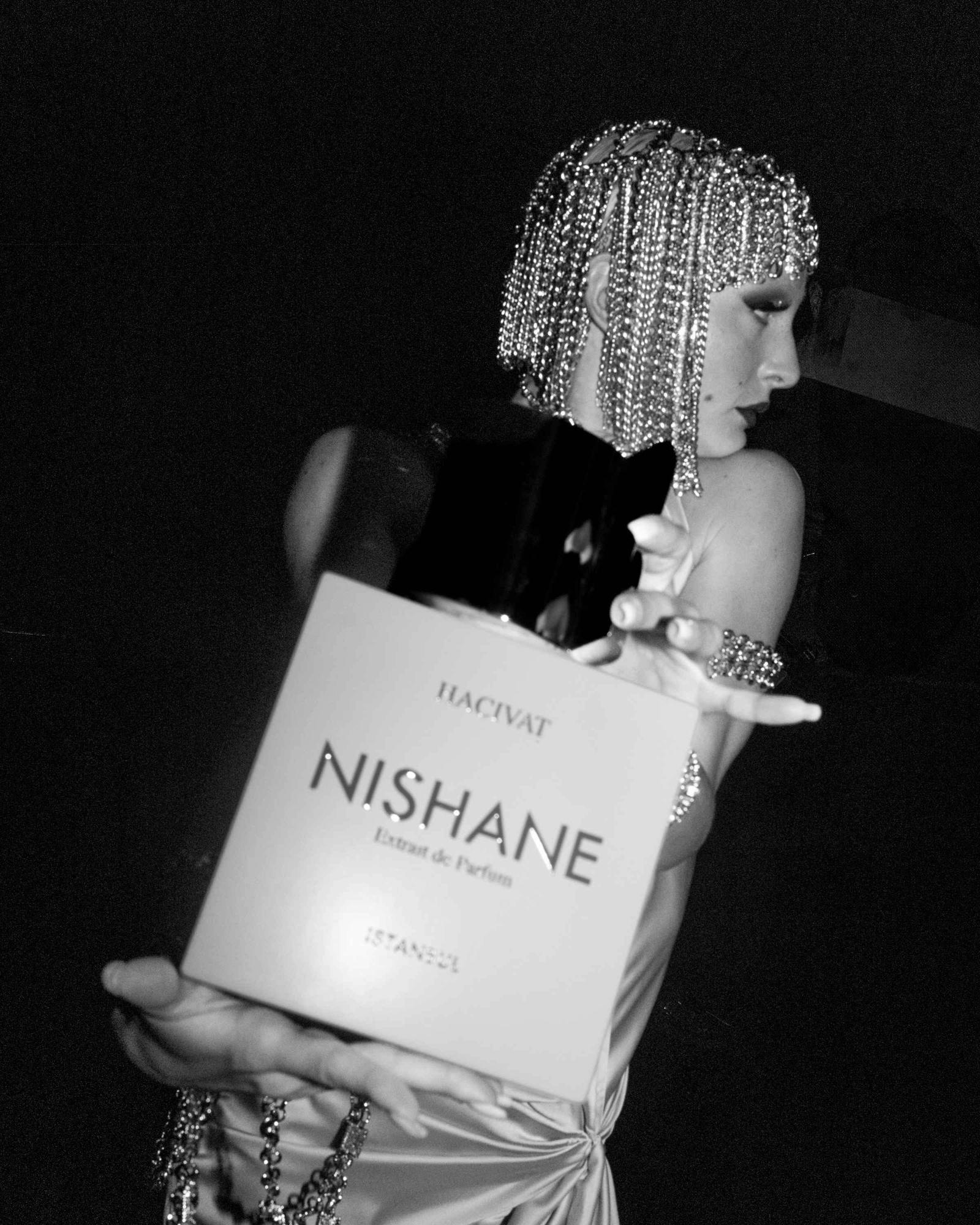 Black and white photo of a model in a beaded silver headpiece and satin dress holding a Nishane Hacivat box
