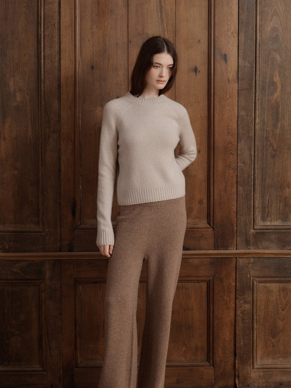 Model wearing a light beige cashmere sweater and taupe trousers in a wood-panel studio