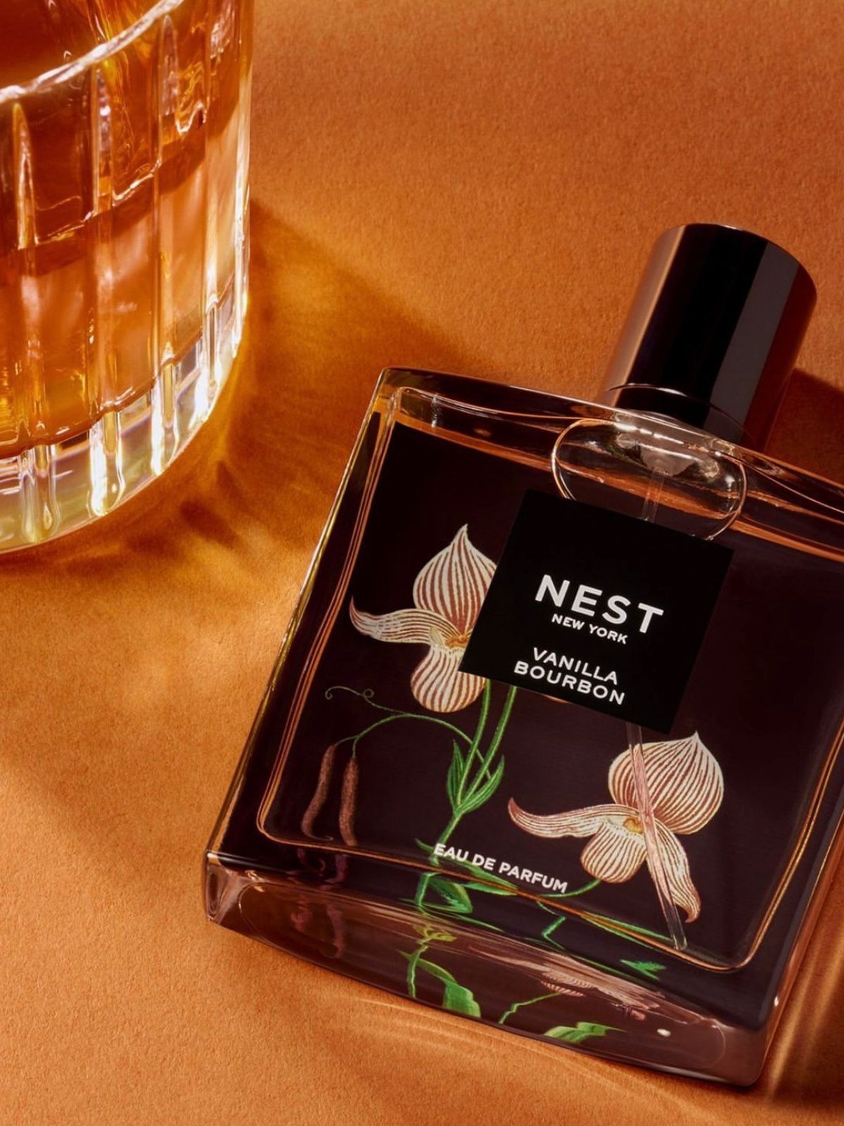 NEST Vanilla Bourbon eau de parfum bottle with orchid illustration lying on amber velvet beside a glass of whiskey