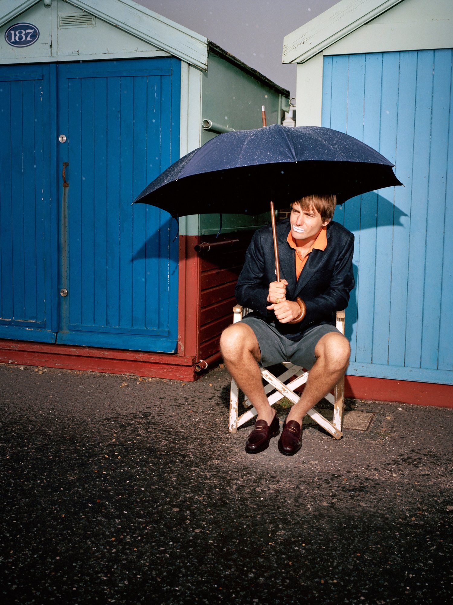 Man under an umbrella on a folding chair in front of blue beach huts